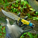 A hand in a black glove holds the Texan Knives Handcrafted 10" Damascus Steel Hunting Knife with a patterned blade, brass guard, and yellow-green-blue resin pakkawood handle. Blurred greenery and wooden fencing form the background.