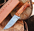 The Texan Knives Premium 10-Inch Damascus Steel Hunting Knife with a rosewood handle and patterned blade rests on stacked logs beside its leather sheath. A perfect gift item, this handmade knife blends elegance and craftsmanship.