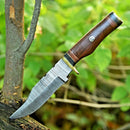 The Texan Knives Damascus Steel Hunting Knife, featuring a 10" blade, brass guard, and resin pakkawood handle, is embedded in a tree stump as its patterned steel gleams with green foliage softly blurred in the background. Leather sheath included.