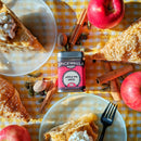 The Spicewalla Apple Pie Spice tin by Spicewalla sits among fresh apples, slices of pie topped with whipped cream, cinnamon sticks, nutmeg, and a fork atop a yellow gingham tablecloth, all glowing in warm sunlight—an ideal autumn setting to showcase this comforting seasonal spice blend.