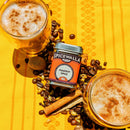 Two frothy drinks dusted with Spicewalla Pumpkin Spice sit next to a tin of Spicewalla’s signature blend, surrounded by coffee beans, cinnamon sticks, and nutmeg on a bright yellow cloth—the perfect inspiration for autumn baking.