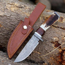 The Texan Knives Damascus Steel Hunting Knife – 10" with a Damascus steel guard and stag horn handle is shown beside its brown leather sheath, both displayed on rugged tree bark.