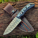 The Texan Knives Custom Handmade 9" Damascus Steel Hunting Knife with a black and blue G10 handle is displayed on its leather sheath, both set atop a tree log with grass in the background.