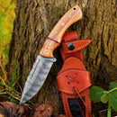 The Texan Knives Custom Handmade 10" Damascus Steel Hunting Knife with Olive Wood Handle and Leather Sheath is displayed against a tree trunk, with the brown sheath resting on the ground beside it.