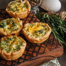Four mini quiches with golden crusts and herb-filled egg centers rest on a wooden board. Fresh rosemary, a white egg, and a jar of The Spice Lab Savory Whole Leaf 1 lb from The Spice Lab are in the background, highlighting the use of premium foodservice herbs that add depth to every bite.