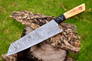 The CUSTOM HANDMADE FORGED DAMASCUS STEEL CHEF KNIFE KITCHEN KNIFE WOOD HANDLE by Texan Knives, featuring a patterned Damascus blade and wood handle, rests on bark atop a tree stump with green grass in the background.
