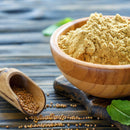 A wooden bowl filled with The Spice Lab Organic Ground Mustard sits on a rustic wooden board alongside a scoop of whole mustard seeds. Some seeds are scattered nearby, with green leaves in the background—a perfect addition for anyone looking to enhance cheese dishes or craft unique spice blends with The Spice Lab’s premium organic product.