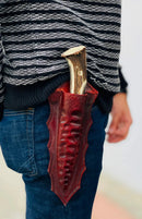 A person wearing blue jeans and a black-and-white striped sweater has a Texan Knives Custom Forged 1095 High Carbon Steel Hunting Knife with a 12” antler horn handle in a leather sheath attached to their belt.