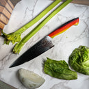 The Spaceman Knives EM Wave Chef Knife by Spaceman Knives, with a patterned Damascus blade and red handle, rests on marble beside celery, iceberg lettuce leaves, and a quarter white onion—a professional chef’s essential tool.