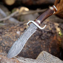 The Texan Knives Damascus Steel Western Bowie Knife—12" with a Damascus steel guard and stag horn handle—rests embedded in a log outdoors, accompanied by its rugged leather sheath, ready for any adventure.