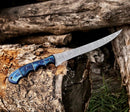 The Texan Knives Handmade 8" Rasp Steel Sheep Horn Hunting Knife, featuring a curved patterned blade and blue ergonomic handle, is displayed outdoors on a log with its leather belt sheath nearby.