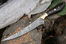 The Texan Knives Damascus Fillet Knife – 13" features a patterned blade, brass bolster, and dark rosewood handle. Resting on a tree stump beside a partially filleted fish, wood shavings surround this premium fishing knife with leather sheath.