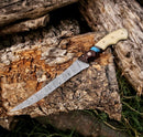 The Texan Knives Fishing Fillet & Boning Knife—13" Damascus steel with a camel bone and rosewood handle—rests on a split log outdoors, surrounded by wooden logs and grass. Includes leather sheath.
