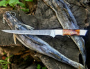 The Texan Knives Custom Handmade Damascus Filleting Knife – 13" Camel Bone Handle with Leather Sheath rests atop two whole, raw fish on a rough tree stump outdoors.