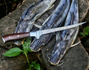 Three catfish with stringers rest on a rough wooden surface outdoors. A Texan Knives Premium Custom Handmade Damascus Steel Fillet Knife – 13” Rosewood Handle lies diagonally across the fish, with green leaves visible nearby.