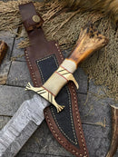 The Texan Knives Custom Handmade Damascus Steel Bowie Knife features a 13-inch patterned blade, bone handle with brass guard, and comes with a stitched leather sheath displayed on stone surrounded by natural elements.