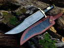 The Texan Knives 13-Inch Damascus Steel Cowboy Bowie Knife with a black buffalo horn handle and leather sheath rests on aged wood, surrounded by green moss—an ideal gift for knife enthusiasts.