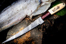 The Texan Knives Custom Handmade Filleting Knife for Fish features a 13" Damascus steel blade and camel bone & rosewood handle, resting on wood beside a half-filleted fish and green leaves.