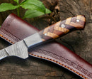 The Texan Knives Handmade Damascus Fillet Knife 8”, featuring a rosewood and olive wood handle, rests on its leather sheath atop a rough tree stump with a green plant in the background.