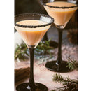 Two martini glasses with creamy cocktails and black salt rims are set on a rustic surface surrounded by pine branches and a pinecone. The striking rims, dusted with The Spice Lab Black Obsidian Salt Cocktail Rimmer, add an elegant touch to this festive scene.