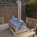 A WPPO LLC Direct WPPO Lil Luigi Portable Oven, constructed from stainless steel, rests atop a wooden patio table. This wood-fired oven includes a tall chimney, wooden handles, and foldable metal legs, all set against the backdrop of a wooden fence and lush green garden hedge.