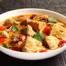 A white bowl filled with spicy fish curry, featuring chunks of fish, red chili peppers, tomatoes, and green curry leaves in an orange-colored sauce enriched with The Spice Lab Organic Ground Turmeric rests on a dark wooden surface—a true celebration of Indian cooking.