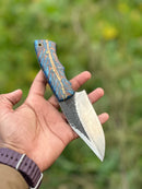 A hand holds the Texan Knives Hunters Knives – a 9" handcrafted D2 steel blade with an epoxy pine cone resin handle and leather sheath, shown against a blurred green outdoor background.