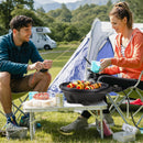 Two people sit by a tent at a campsite, smiling and preparing skewers over the DirectDeals 1350 W Outdoor Electric BBQ Grill with Removable Stand Easy to Install on a small table. Food, drinks, and camping gear surround them.