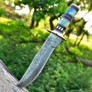 The Texan Knives 13" Damascus Steel Antique Bowie Knife, featuring a brass guard and color bone & pakkawood handle with a leather sheath, is displayed outdoors in wood with blurred green foliage and trees in the background.