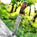 The Texan Knives Antique 13" Damascus Steel Bowie Knife, featuring a hand-forged blade, brass guard, and Pakkawood handle with leather sheath, is shown embedded in a weathered log against a blurred green forest backdrop.
