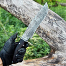 A gloved hand holds the Texan Knives Damascus Steel Antique Bowie Knife for Sale – 13" with a Damascus steel guard and rosewood handle, outdoors by a fallen tree, its included leather sheath resting nearby.