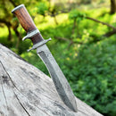 The Texan Knives Custom Handmade 13" Damascus Steel Civil War Bowie Knife with a Rosewood Handle and Leather Sheath is embedded in a weathered tree trunk, set against a blurred, green forest background.