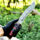 A person in a black tactical glove holds the Texan Knives Antique 13" Damascus Steel Bowie Knife with a brass guard and pakkawood handle, outdoors amidst green foliage.