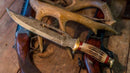 A Texan Knives Damascus Steel Vintage Bowie Knife – 13" with brass guard and stag horn handle, complete with a leather sheath, lies on a wooden table beside a rifle and deer antlers.