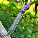 The Texan Knives Vintage Original Damascus Steel Bowie Knife – 13" with a brass guard and camel bone handle is embedded in a tree trunk, set against a blurred green forest background.