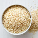 A white bowl filled with The Spice Lab Organic Hulled Sesame Seeds sits on a light wooden surface, with some seeds scattered around the bowl—perfect for adding a nutty crunch to baking or stir-fries.