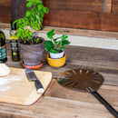 A kitchen scene featuring potted plants, olive oil bottles, a cutting board with dough and a knife, and the WPPO 9" Round Aluminum Pro Turning Peel by WPPO LLC Direct with its long handle resting on a rustic wooden countertop.