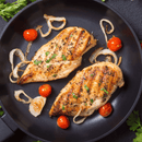 Two hand-trimmed, boneless skinless chicken breasts from the Dude Food Chicks Meat Box by Dude Food are grilled to perfection in a skillet with sliced onions and cherry tomatoes, then garnished with chopped herbs. The natural chicken is well-browned and arranged neatly on a dark surface.