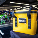 A bright yellow Bob The Cooler Co. 14L "Sidekick" Flip Top Cooler Bag with black straps sits on a table in a garage, displaying its advanced cooling technology from Bob - The Cooler Co., while a green motorcycle and people are blurred in the background.
