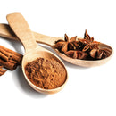 A wooden spoon filled with ground cinnamon and another with The Spice Lab Ground Star Anise by The Spice Lab—both aromatic spices—rest alongside cinnamon sticks on a white background. This arrangement beautifully highlights the warm spice of cinnamon and the licorice-like flavor of The Spice Lab Ground Star Anise in perfect harmony.