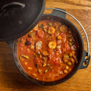 A cast iron pot filled with chunky vegetable stew in tomato sauce sits on a wooden surface, its lid partially open. The rich aroma of The Spice Lab Organic Chili Powder infuses the air, promising bold and vibrant Mexican-inspired flavors in every bite.