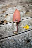 A Texan Knives Damascus Steel 1800s Bowie Knife – 15" with Damascus steel guard and stag horn handle sits in its leather sheath on weathered wooden boards beside a single yellow maple leaf.
