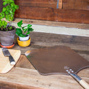 On a rustic wooden table rest the WPPO Traditional Aluminum Long Handle Pizza Peel (16"W x 40"L) by WPPO LLC Direct, a cutting board with a knife, and two potted plants—perfect companions for wood-fired cooking.
