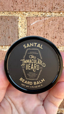 A hand holds a round tin labeled The Immaculate Beard Limited Edition Beard Balm from The Immaculate Beard, featuring gold text on a black lid and infused with nourishing shea butter and sandalwood, set against a brick wall background.