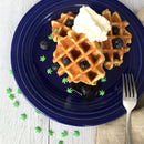 Two waffles topped with whipped cream, blueberries, syrup, and The Spice Lab Hemp Leaf Sprinkles CBD & THC Free—edible party decorations from The Spice Lab—on a blue plate. A fork rests on a beige napkin beside the plate.