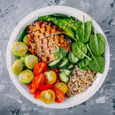 A white bowl filled with grilled chicken, quinoa, cherry tomatoes, Brussels sprouts, spinach, cucumber, and avocado slices sits on a gray textured surface, lightly seasoned with The Spice Lab Organic Ground Coriander—perfect for those who appreciate spice blends often found in Indian cooking.