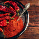 A vintage spoon filled with The Spice Lab Organic Cayenne Pepper rests on a dark plate next to several dried red chili peppers atop a rustic wooden table—perfect for adding authentic heat to Mexican dishes.