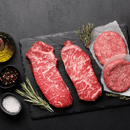 Two raw marbled Kansas City Strip Steaks and two raw American Ground Beef hamburger patties from the Dude Food Common Man Meat Box by Dude Food rest on a black slate, surrounded by rosemary, thyme, coarse salt, peppercorns, and a bottle of olive oil—all set against a dark textured background.