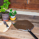 A WPPO 12" Professional Aluminum Pizza Peel by WPPO LLC Direct sits on a rustic wooden counter along with a knife, a wooden cutting board dusted with flour, and two potted plants.