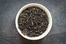 A white bowl filled with loose, dried World Spice Jorhat Assam tea leaves from World Spice sits on a dark, speckled surface.